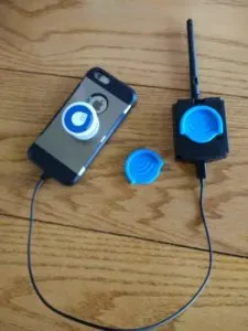Smartphone connected by cable to a small device with an antenna, both displaying matching blue‑and‑white circular patterns, arranged on a wooden surface.