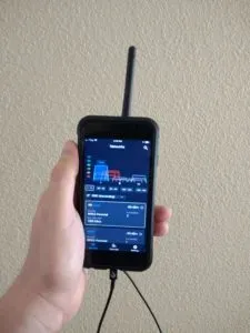 Hand holding a smartphone with an attached antenna, showing an SDR++ interface tuned to 1090 MHz with a live frequency spectrum and signal readouts.