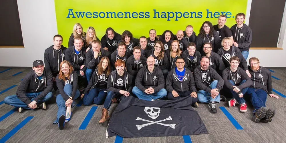 Group photo of the MetaGeek team posing indoors in front of a green and blue “Awesomeness happens here” banner, with many members wearing skull‑and‑crossbones hoodies and a pirate‑themed flag displayed at the front.